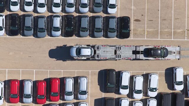 High-angle drone footage showing a car carrier truck navigating a vast parking area filled with rows of brand new vans, showcasing automotive logistics and mass vehicle distribution