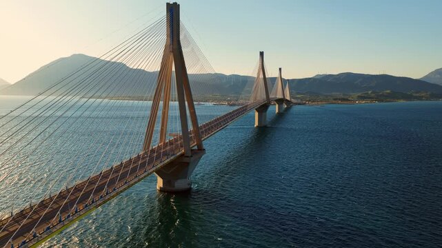 Golden hour aerial footage showing the magnificent rio antirrio bridge, a stunning feat of engineering, connecting the peloponnese peninsula to mainland greece over the deep blue sea