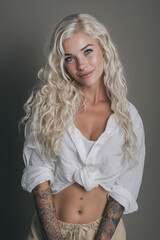 Naklejka premium Young woman with long curly blonde hair and tattoos wearing a white shirt and light pants poses with a smile against a neutral background
