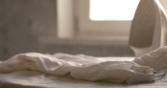 Cinematic close-up iron on table with white sweater. Water drops fall as a spray bottle sprays the sweater before ironing. Window and home interior blurred in background. Soft light and soft image. 4K
