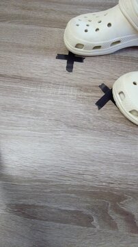 Shoe approaching tape marker on floor, crocs toe hovering over black tape cross, wood laminate surface, directorstyle blocking demonstration, measurement for camera position,
