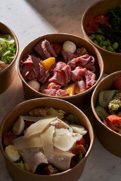 Variety of gourmet salad bowls in craft paper packaging on a white marble surface. Assortment of healthy takeout meals including roast beef, salmon, and chicken.