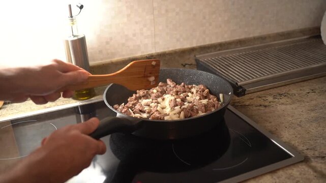 Male hand stirring chopped meat and onion with a wooden spatula in a hot pan on an induction cooktop