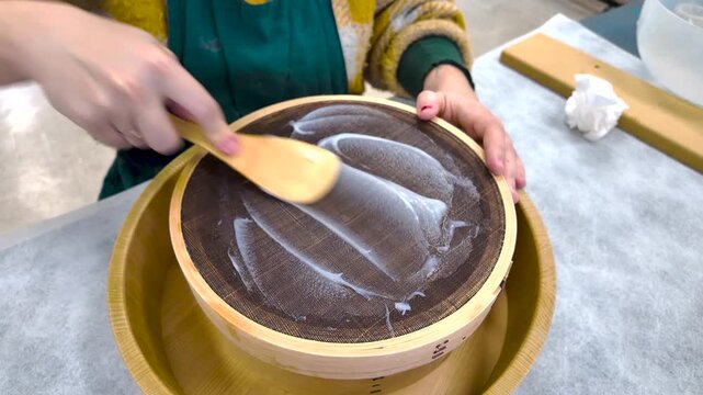 Mains d'une femme faisant passer une pr&eacute;paration de colle avec une spatule en bois dans un tamis afin d'&eacute;liminer les grumeaux. Geste artisanal, r&eacute;p&eacute;titif et m&eacute;ticuleux.