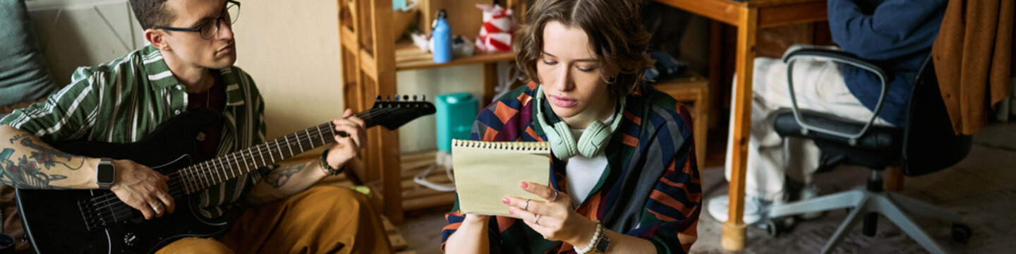 Header of young musicians rehearsing at home, man playing electric guitar, woman writing lyrics in notebook, showing songwriting teamwork. Ideal for music education, creative process, band promotion
