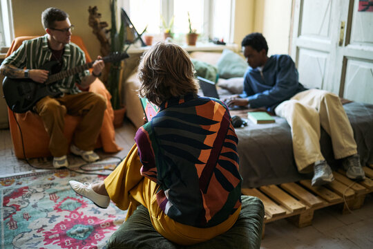 Young roommates collaborating in shared apartment, playing guitar and using laptop while relaxing together, showing creative lifestyle. Useful for youth culture, rental living, community marketing