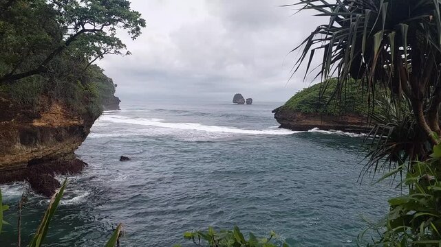 Scenic tropical coastal bay featuring turquoise ocean water, rocky island formations, and lush green cliffs under cloudy skies at Tanjung Penyu beach, East Java, Indonesia.