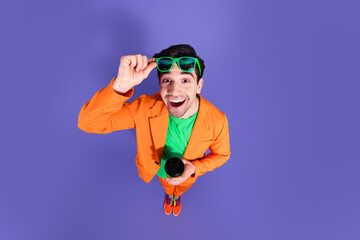 Naklejka premium Excited young man in orange suit and green sunglasses smiling and holding a coffee cup on a purple background