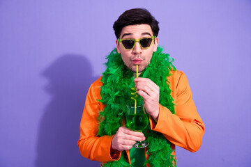 Naklejka premium Man in orange jacket and green feather boa sipping a green cocktail through a straw while wearing sunglasses on purple background, festive party and playful celebration portrait