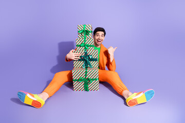Naklejka premium Happy man in bright orange suit holding stacked Christmas gifts on purple background with colorful sneakers for a playful festive celebration