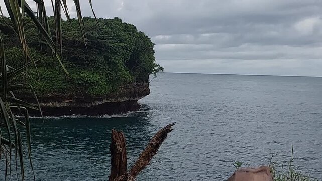 Beautiful small tropical island covered in green vegetation surrounded by calm turquoise ocean water under dramatic cloudy sky at Tanjung Penyu beach, East Java, Indonesia.