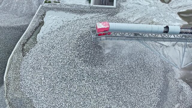 A truck unloads gravel at a construction site while snow covers the ground. Equipment moves material to a large pile for future use. The area shows a working environment in winter.