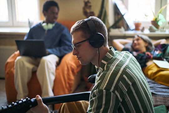 Young adult Caucasian man wearing headphones and playing guitar at home while friends using laptop and relaxing nearby, showing creative leisure and shared living. Ideal for youth lifestyle marketing