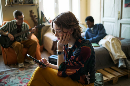 Young adult Caucasian woman checking smartphone while sitting with friends at home, showing digital distraction and social disconnect. Useful for screen time or youth lifestyle marketing