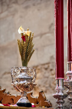 Ornate silver vessel with wheat and flowers beside tall red candles on a processional platform, reflecting Catholic symbolism of devotion and liturgical tradition