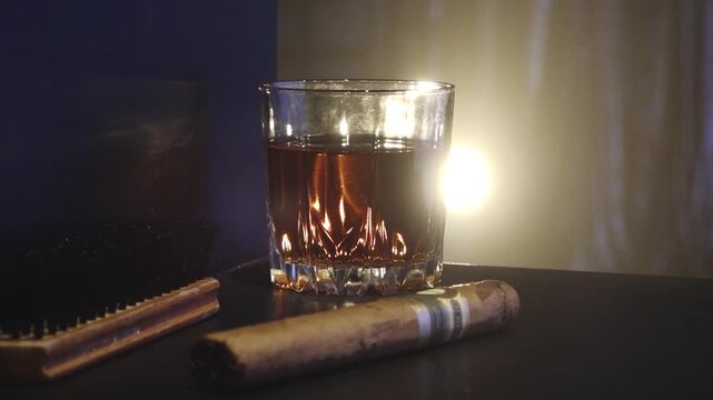 Low-lit still life of amber liquor in a glass beside a cigar and comb, with warm backlight, shadows, and copy space