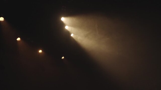 Bright stage spotlights cut through smoky darkness, forming warm beams across a minimalist scene with ample copy space