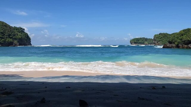 Turquoise ocean waves crashing on sandy beach with small tropical island view, located at Gatra beach, East Java, Indonesia.