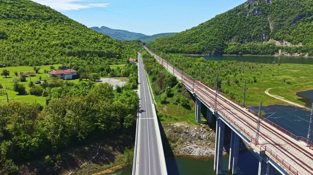 A highway runs parallel to train tracks that cross a lush valley near a river. The landscape includes hills and a small house near the road. It is daytime with clear skies.
