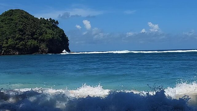Turquoise ocean waves crashing on sandy beach with small tropical island view, located at Gatra beach, East Java, Indonesia.