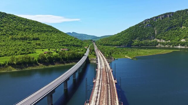 A highway runs parallel to a railway line, both crossing over a river. Lush green hills rise in the background under a clear blue sky. The scene shows nature and infrastructure in harmony.