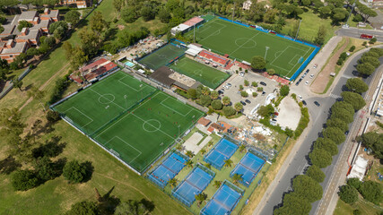 Aerial drone view of a large multi-sport complex featuring several green soccer pitches and blue padel courts, surrounded by lush parkland and urban infrastructure on a bright, sunny day. © Stefano Tammaro