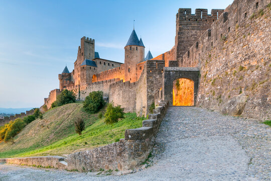 Historic medieval fortified city walls and towers illuminated at dusk, with a cobblestone path leading to an archway, a prime tourism destination in Carcassonne, Languedoc-Roussillon, France.