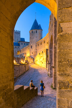 Travelers framed by an archway explore the illuminated medieval city of Carcassonne at dusk, a historic tourism destination in Languedoc-Roussillon, France.