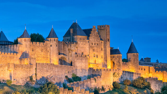 Illuminated medieval city walls and towers stand against a deep blue dusk sky, showcasing European history and a popular travel destination in Carcassonne, Languedoc-Roussillon, France.