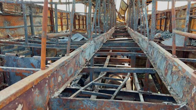 Tracking view through charred sleeper bus floor structure with exposed metal framework, rusted surfaces and severe fire destruction inside damaged vehicle!