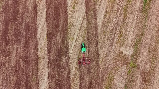 A tractor is driving on a brown field. It is pulling a plow as it works the soil. The environment is open and flat with no buildings nearby. The activity takes place during daylight.