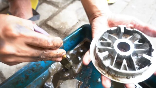 The mechanic is cleaning the spare part of an automatic motorbike, the roller housing, using a brush manually moistened with gasoline to remove the dirt that is stuck to it.
