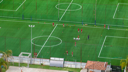 High-angle shot of players in red uniforms training on a vibrant green artificial turf soccer field with white markings and floodlights, showcasing a professional sports facility layout. © Stefano Tammaro