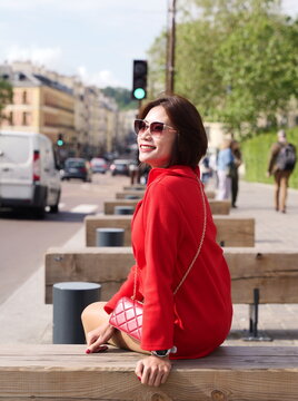 A woman in a red jacket and sunglasses sits on a wooden bench looking toward a city street
