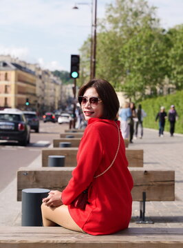 A woman in a red jacket and sunglasses sits on a wooden bench looking toward a city street