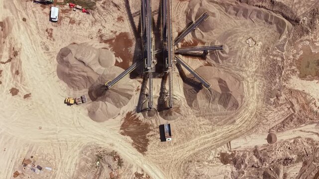 Dust and machinery are present in a mining area where sand and gravel are being processed. Equipment moves materials to various locations. Trucks and piles are visible in the background.