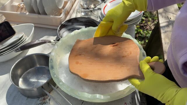 Woman in yellow gloves washing dishes in basin outdoors person scrubs large wooden cutting board with sponge under a kitchen faucet. Bright lighting, close-up shot, steady camera, clean environment
