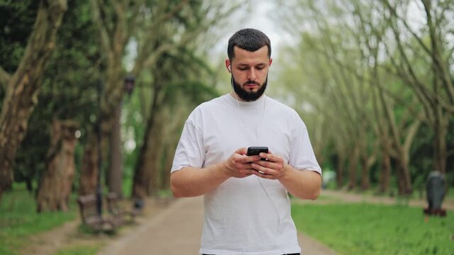 Young man standing on park alley holding phone and wearing wired earbuds outdoors. Person listening to music, podcast or checking navigation. Lifestyle of modern male in nature environment.