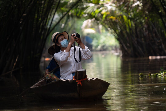 Young Asian woman tourist wearing a face mask riding a traditional wooden boat taking photos with a camera in a tropical canal tunnel. New normal safe travel and eco tourism concept.