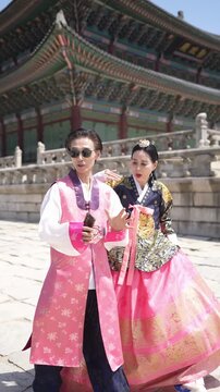Two people in traditional Korean attire stand before a historic palace. One wears a pink hanbok with floral patterns. The other wears a dark, ornate hanbok with pink trim