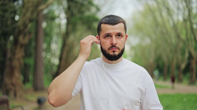 Young man putting on white wired earbuds before starting outdoor training session in park. Person prepares for fitness workout, jogging or morning exercise in nature. Active lifestyle concept.