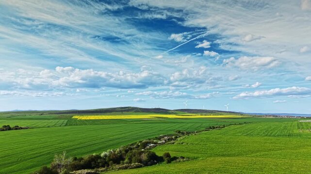 Fields of green grass cover the landscape with yellow patches in bloom. Wind turbines rotate gently in the distance as clouds fill the blue sky overhead.