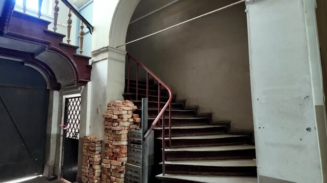 Interior of an old tenement house staircase featuring wooden steps, metal handrail, stack of bricks on the floor, mailboxes, historic architectural arches, natural side lighting, and raw plastered wal
