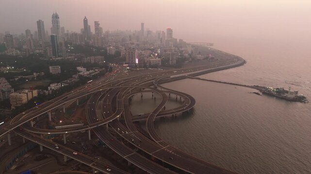 Aerial view of the Coastal Road interchange with complex highway loops and the city skyline under a hazy sunset in Mumbai, Maharashtra, India.