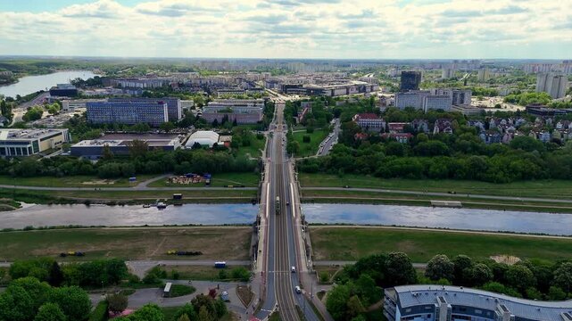 Aerial panoramic view of Saint Roch Bridge spanning the Warta River in Poznan, urban landscape featuring Poznan University of Technology buildings, green riverside embankments, blue sky with white clo