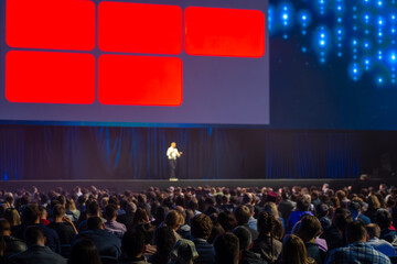 Conference speaker on stage addressing large audience during business presentation, bright screen...