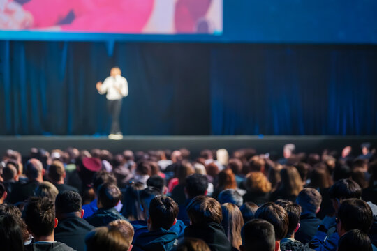 Large audience facing blurred presenter on stage during conference, focused atmosphere, professional event, public speaking, live presentation.