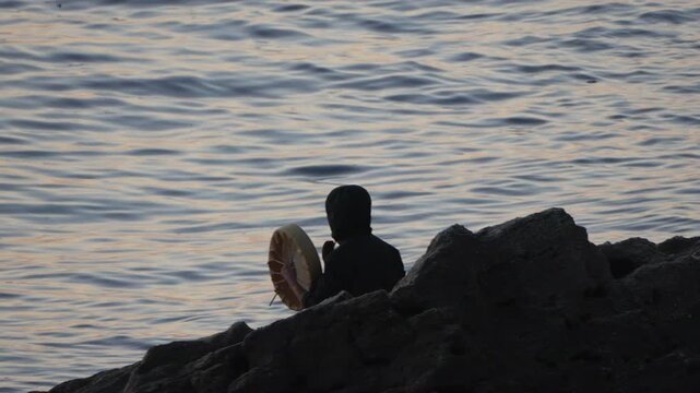 Shaman drumming ocean, hooded figure performs ancient ritual on rocky coast during peaceful sunset.