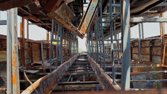Tracking shot through the charred interior of a fire destroyed sleeper bus showing exposed metal frames, collapsed flooring and skeletal structure