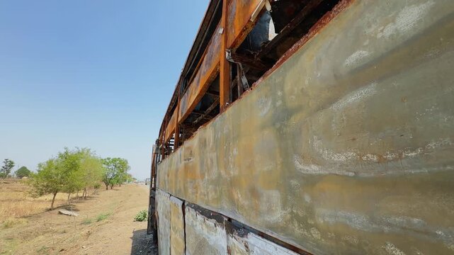 Tracking side shot of a fire destroyed sleeper bus showing scorched metal body panels, shattered window frames and rusted exterior beside a rural roadside!